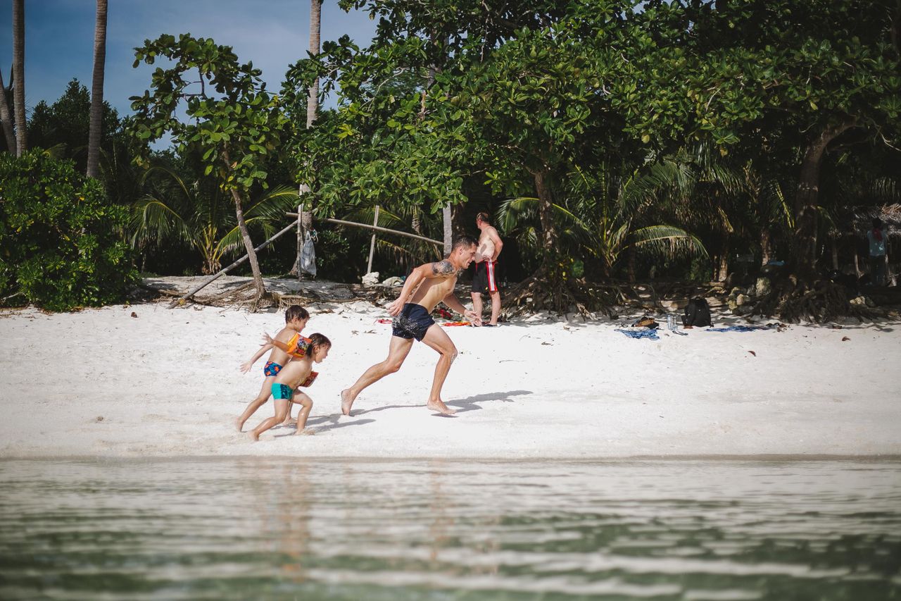 A parent and two children playing and running on a palm-backed sandy beach
