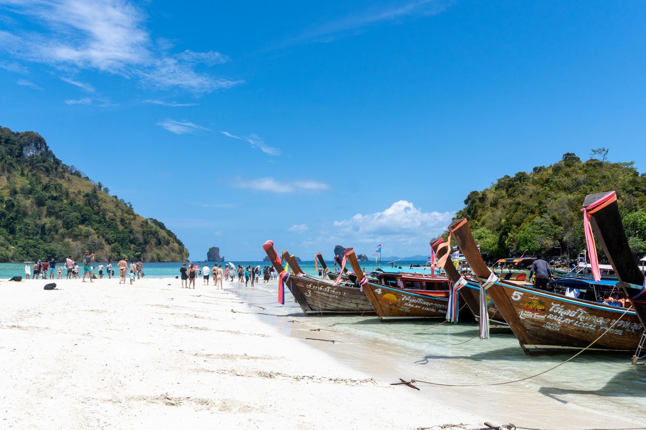 Traditional Thai longtail boats lined up on a busy Andaman Sea beach with limestone islands behind